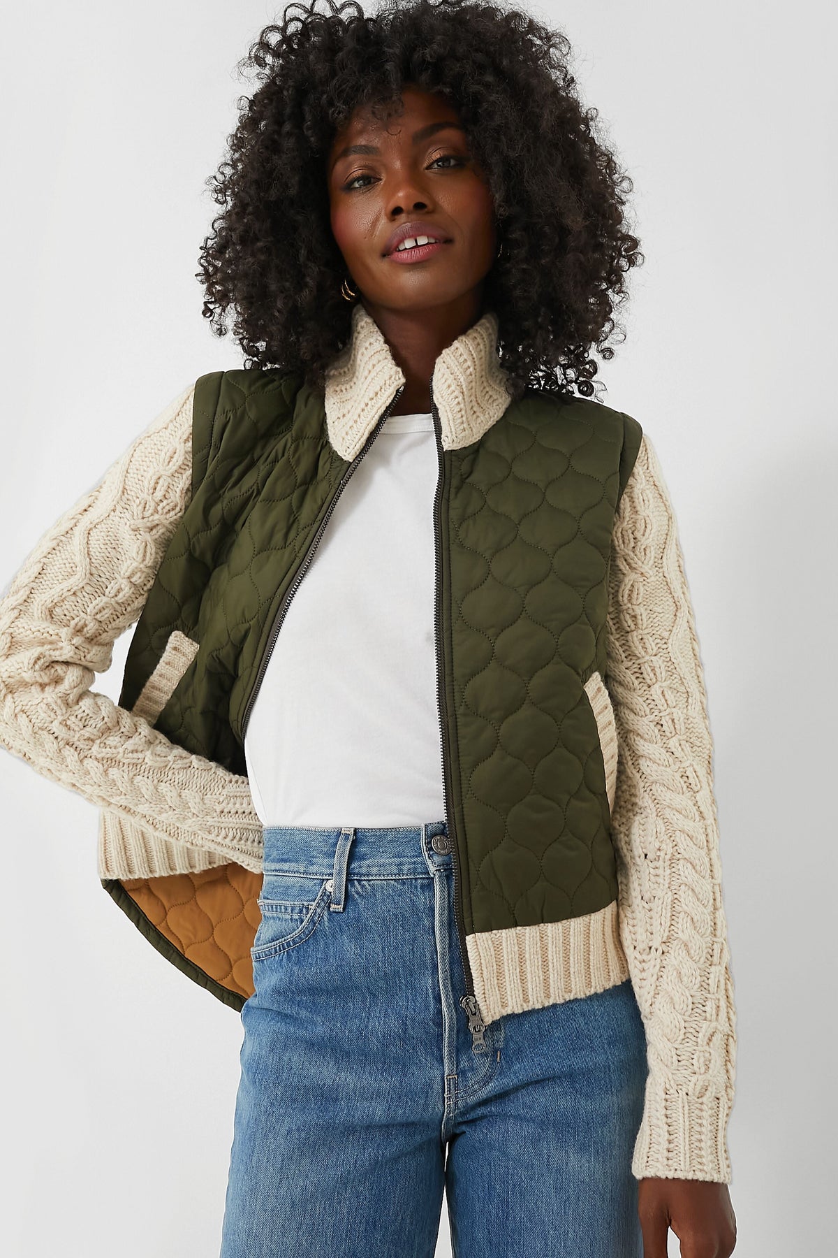 A woman wears a green quilted Veronica Beard Patra Jacket with cream knit sleeves, paired with a white t-shirt and blue jeans.